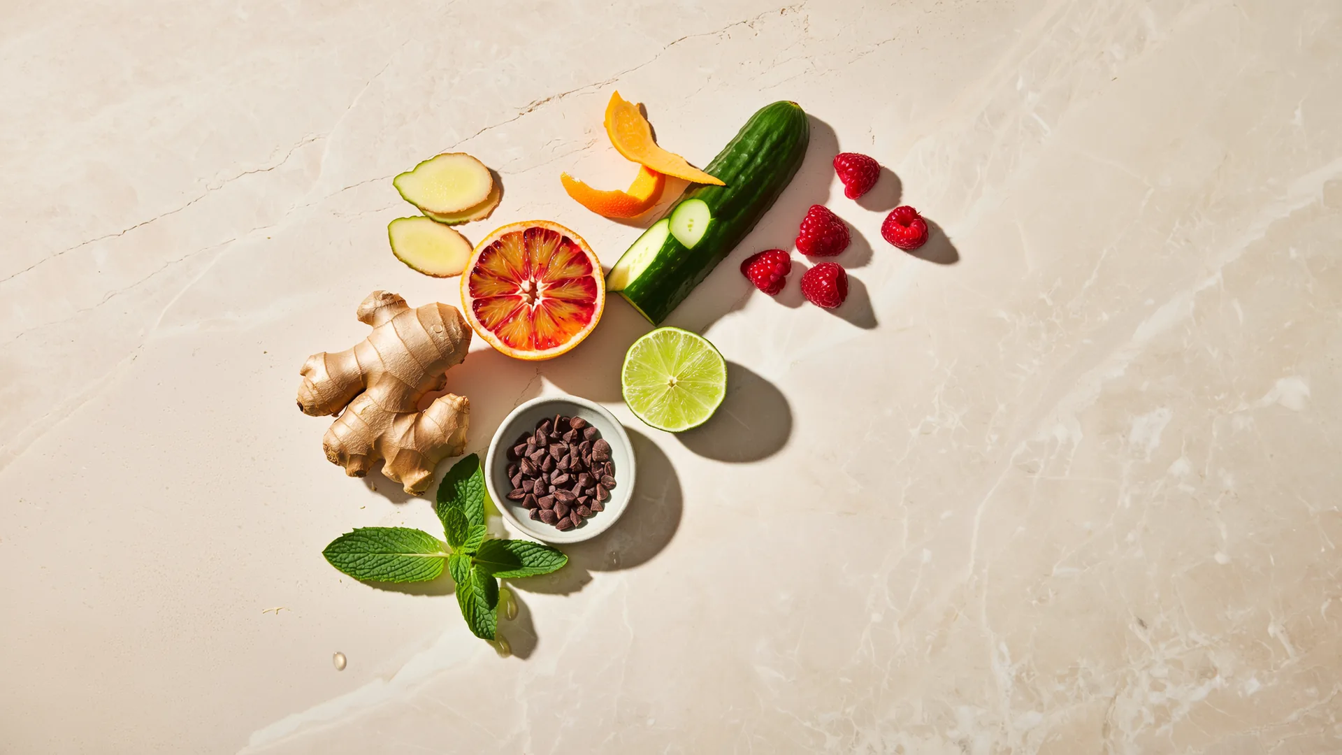 Fresh ginger, citrus, cucumber, herbs, cacao nibs and raspberries on a stone surface.